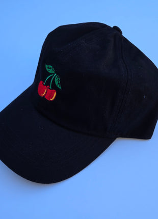 A black baseball cap featuring an embroidered cherry design on the front, set against a solid blue background.
