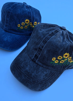 Two dark blue washed baseball caps with embroidered yellow sunflowers on the side, displayed on a solid blue background.