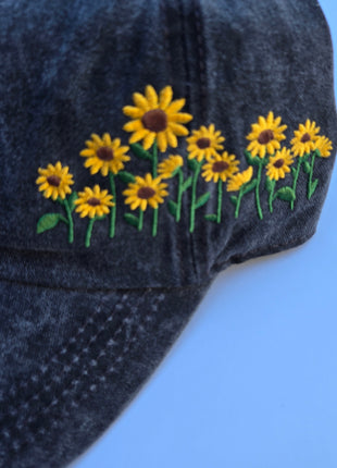 A close-up of a dark washed denim baseball cap featuring embroidered yellow sunflowers with green stems on the side, set against a solid light blue background.