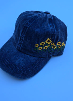 A dark blue washed cotton baseball cap featuring embroidered yellow daisies with green stems on the side, set against a solid blue background.