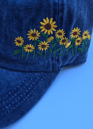 A close-up of a dark blue denim cap featuring embroidered yellow sunflowers with green stems on the side, set against a solid blue background.